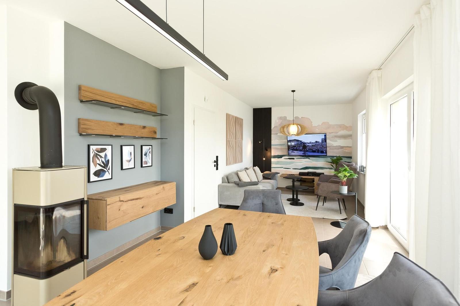 Essbereich mit Holztisch und grauen Stühlen. Im Hintergrund Wohnzimmer mit Sofa und Fernseher.