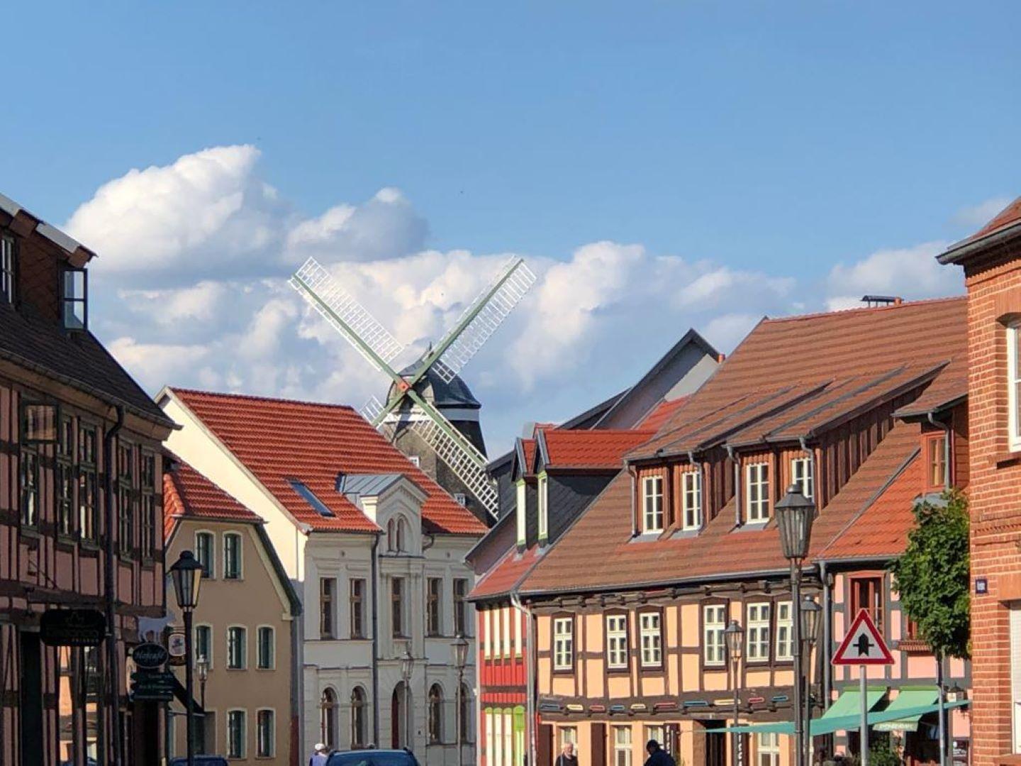 Straßenszene mit Fachwerkhäusern und Windmühle im Hintergrund unter blauem Himmel.