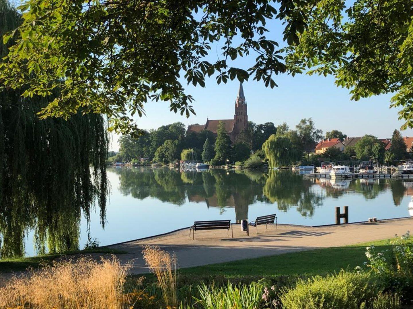 Seeufer mit Bänken, Kirche und Booten im Hintergrund.