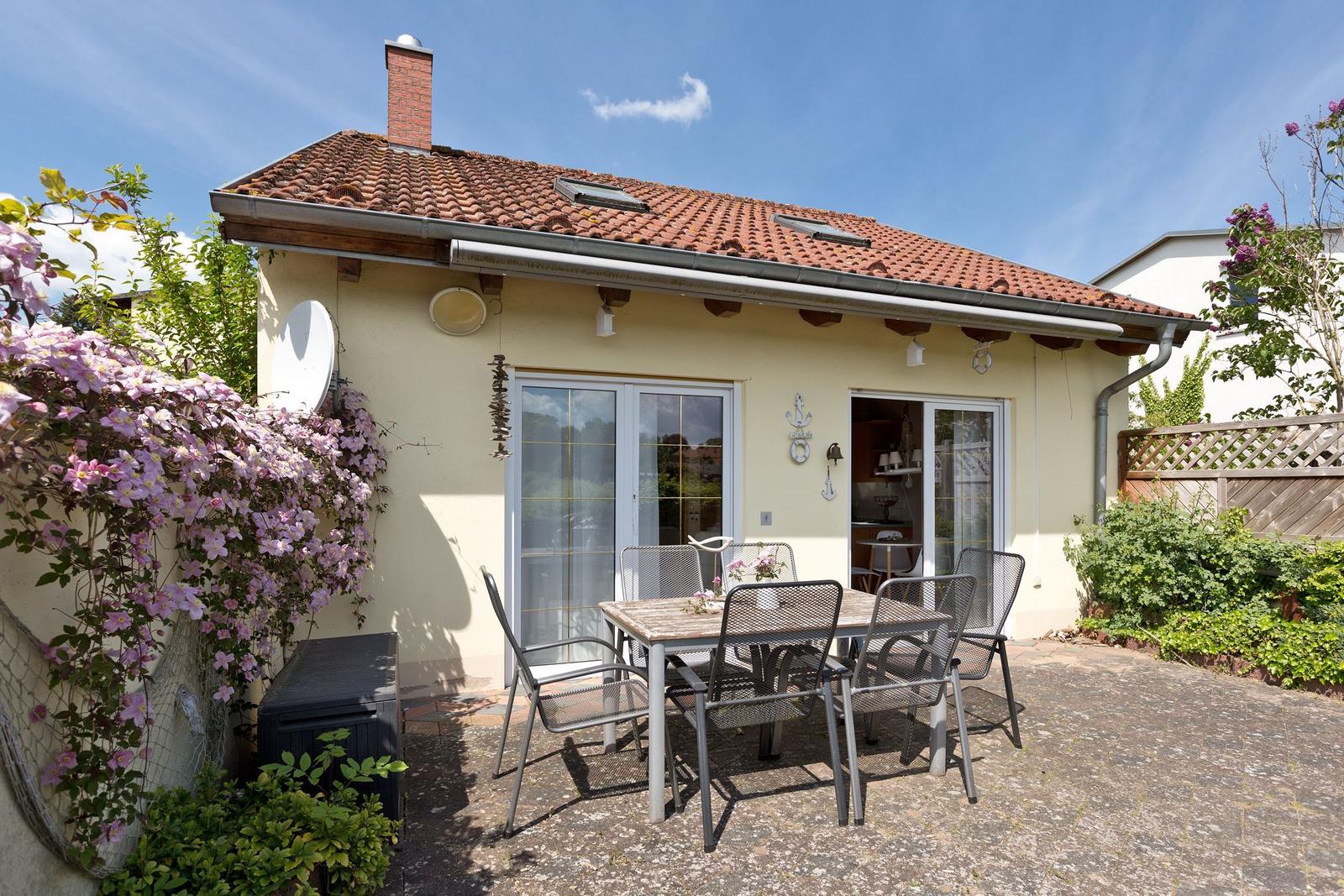 Haus mit Terrasse, Garten und Tisch mit Stühlen. Dach mit Schornstein und Fenstern.