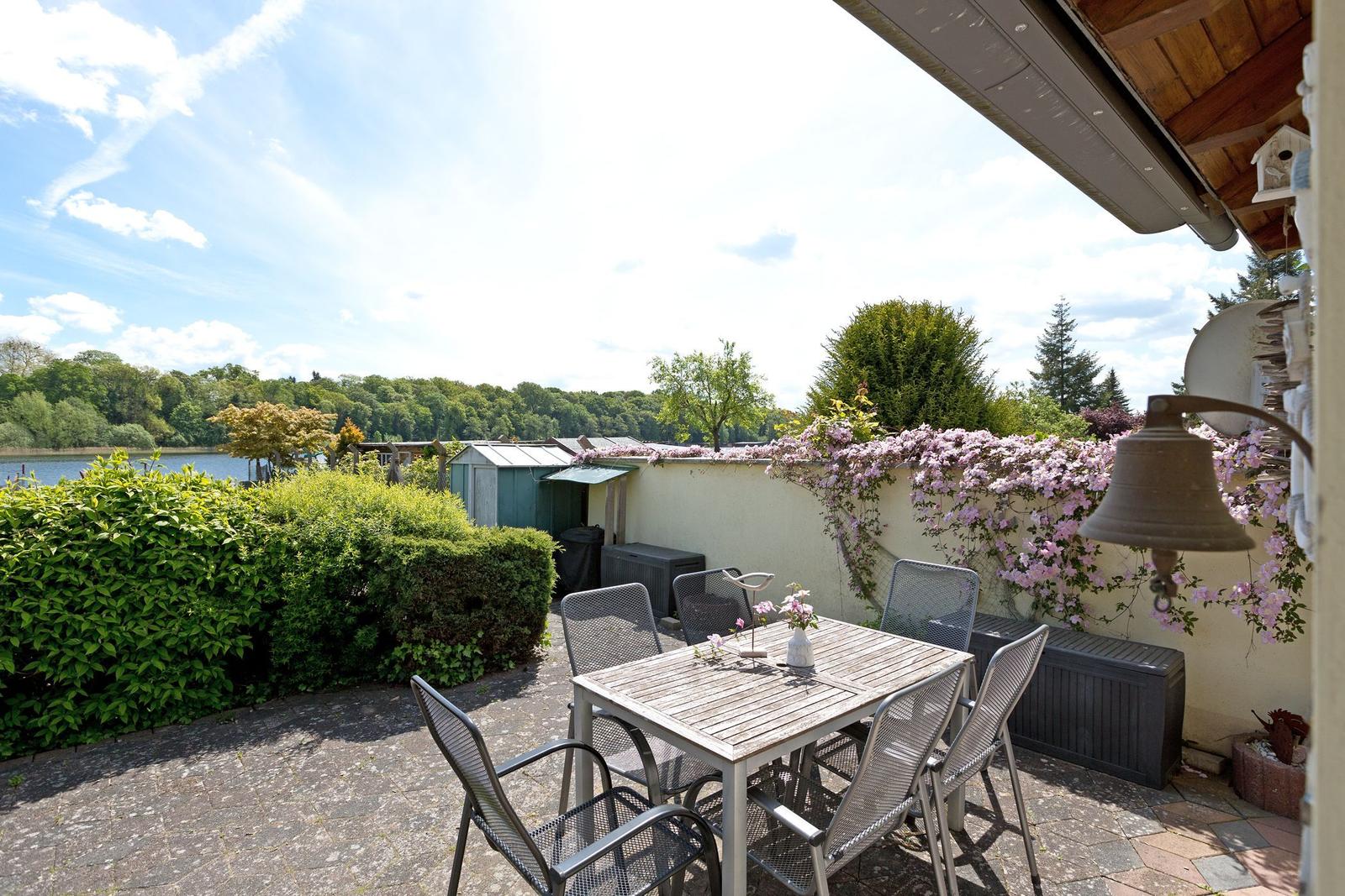 Terrasse mit Tisch und Stühlen, Blick auf einen See und grüne Bäume.