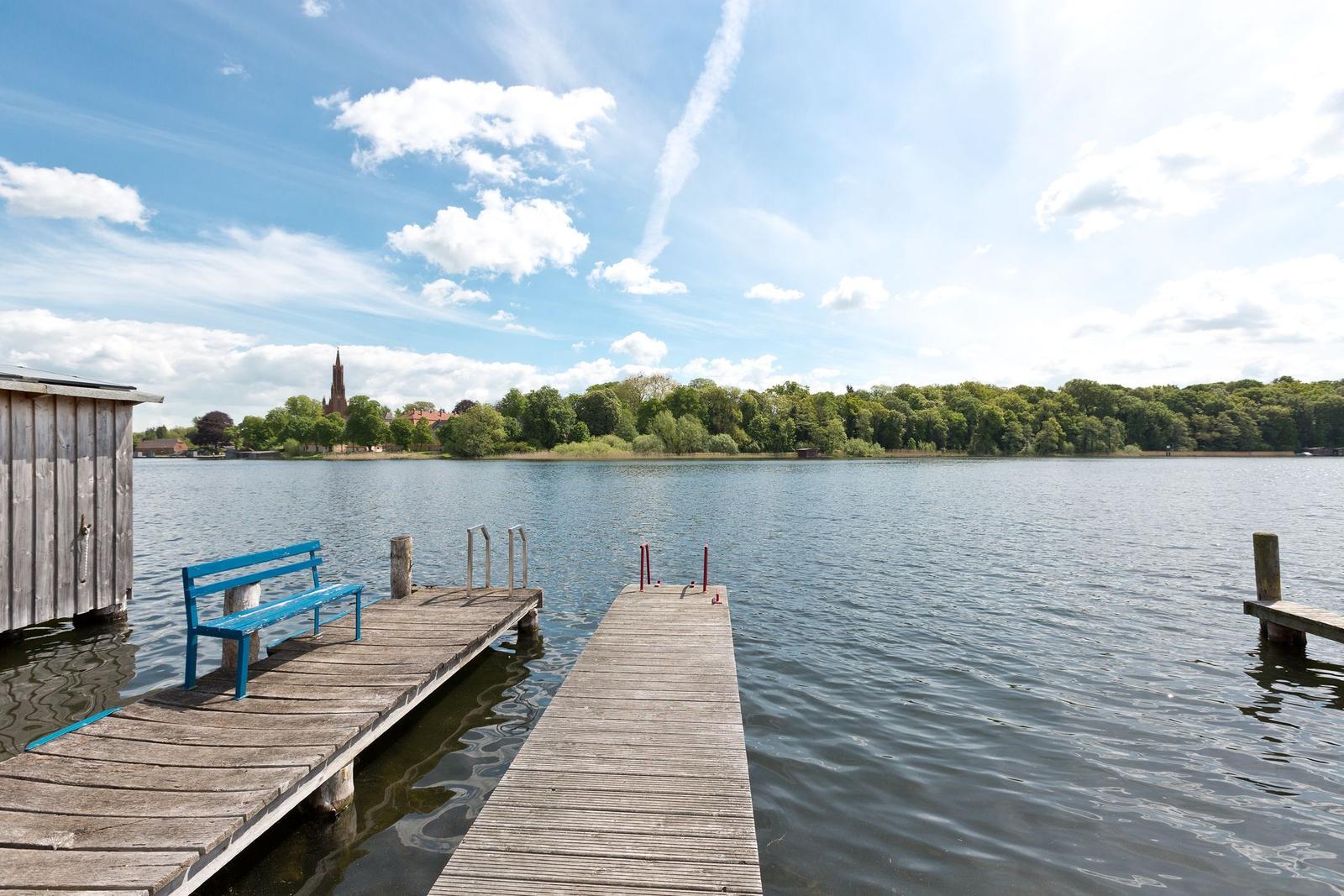 Holzsteg mit blauer Bank am Seeufer. Im Hintergrund Wald und Kirche.