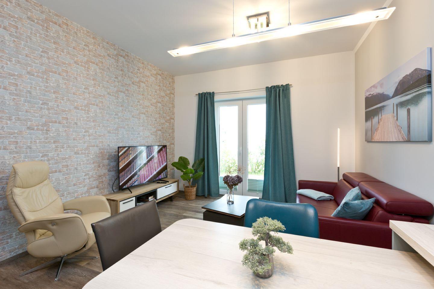 Wohn- und Essbereich mit Sofa, TV und Essplatz. Wand aus Ziegelsteinen und Fenster mit Vorhängen.