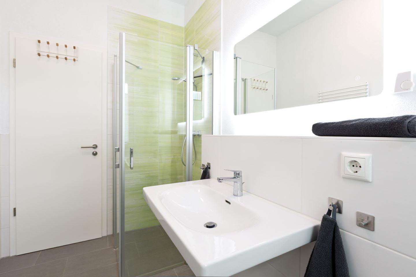 Badezimmer mit Dusche, Waschbecken und Spiegel. Wandfliesen in Grün und Weiß.