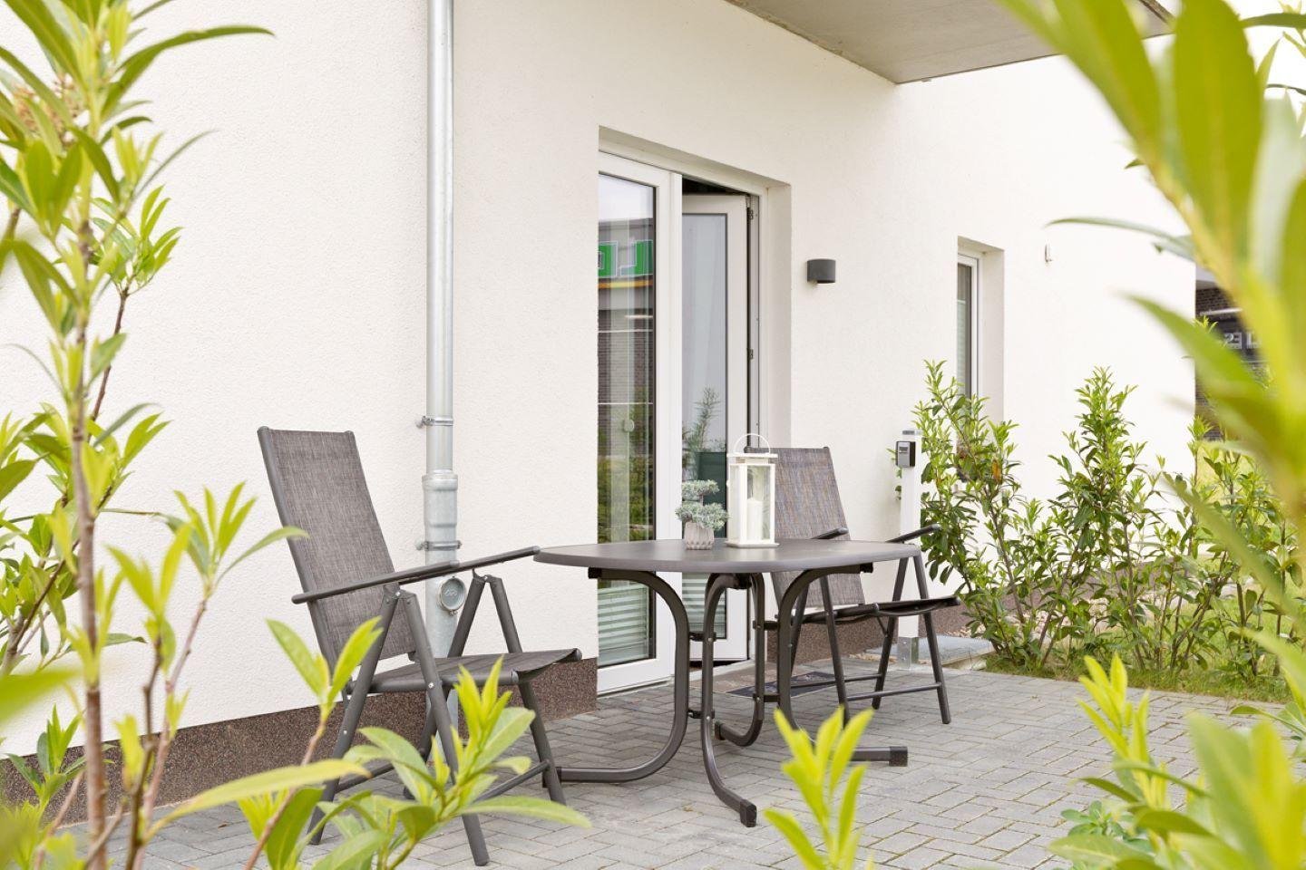 Terrasse mit Tisch und zwei Stühlen, umgeben von Pflanzen und einer Glastür.