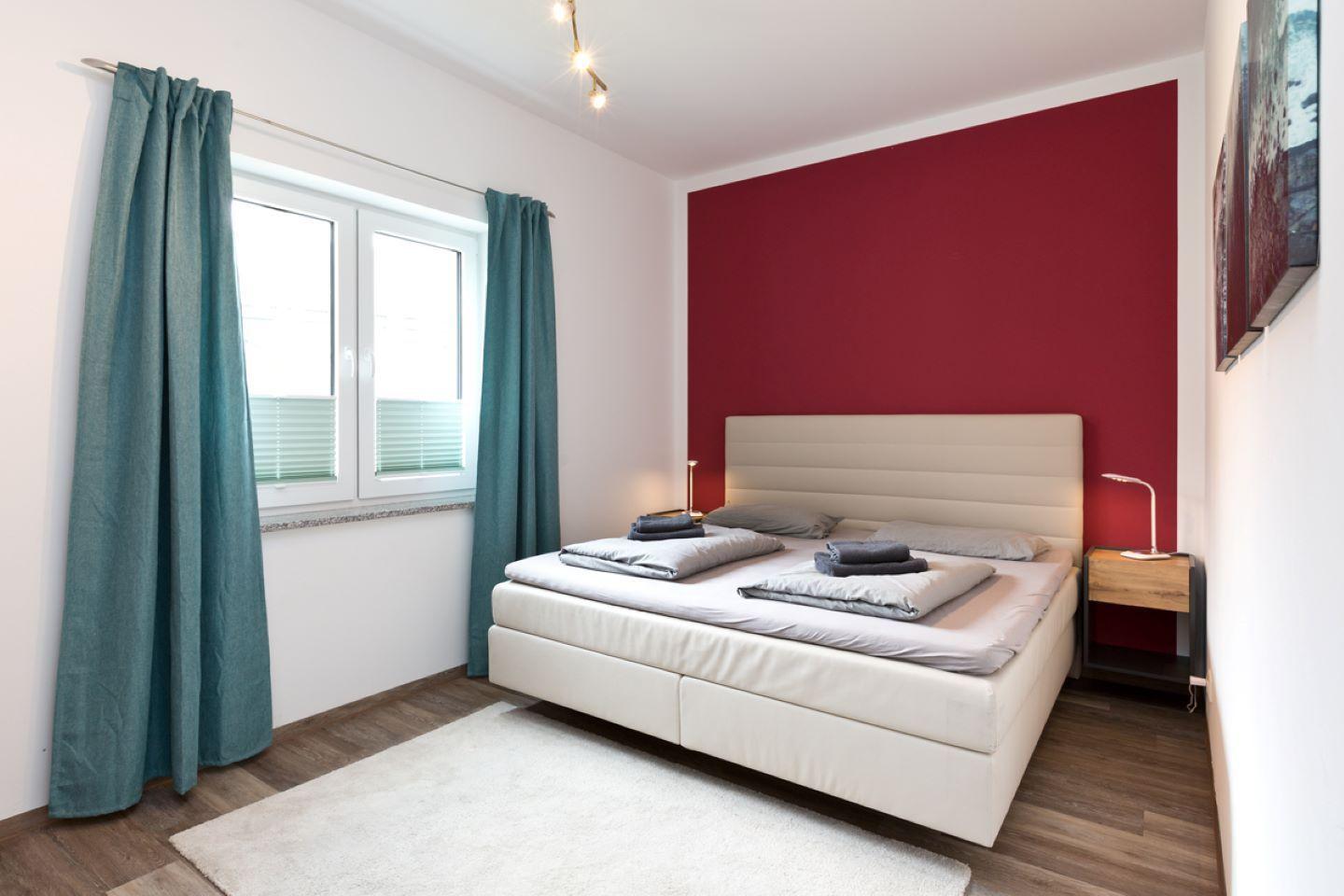 Schlafzimmer mit weißem Bett, roter Wand und blauen Vorhängen.