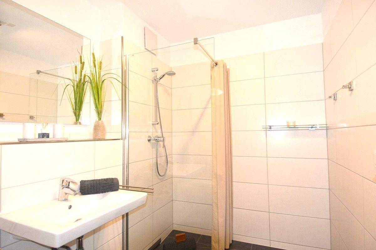 Badezimmer mit Dusche, Waschbecken und Spiegel. Weiße Fliesen und Pflanze sichtbar.