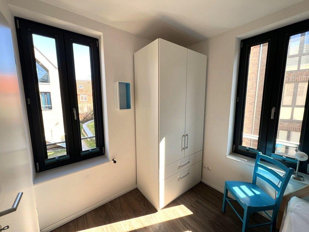Schlafzimmer mit weißem Schrank, blauer Stuhl und zwei Fenstern mit Blick auf Gebäude.