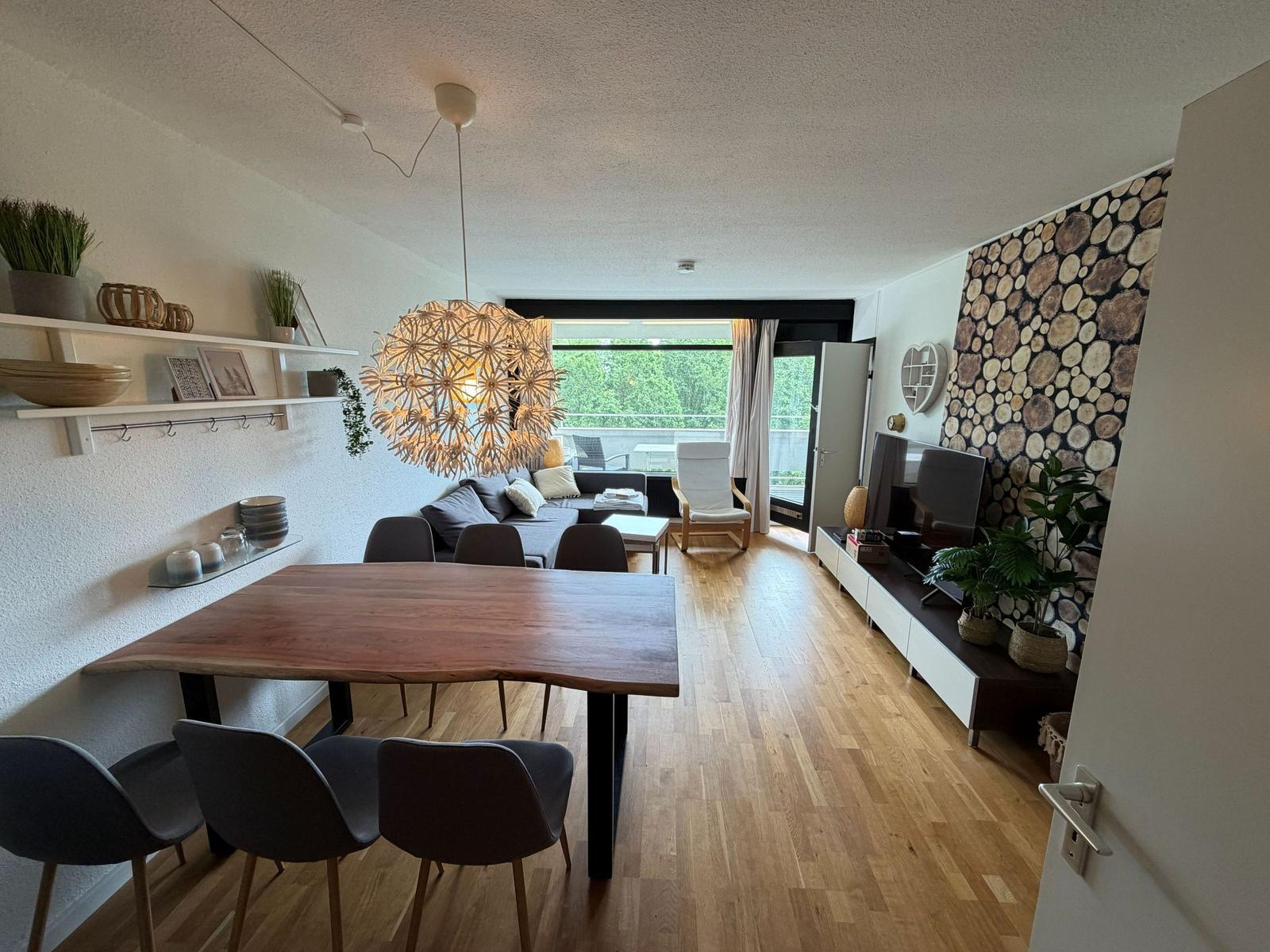 Essbereich mit Holztisch und Stühlen. Wohnbereich mit Sofa, Fernseher und Holzwand.