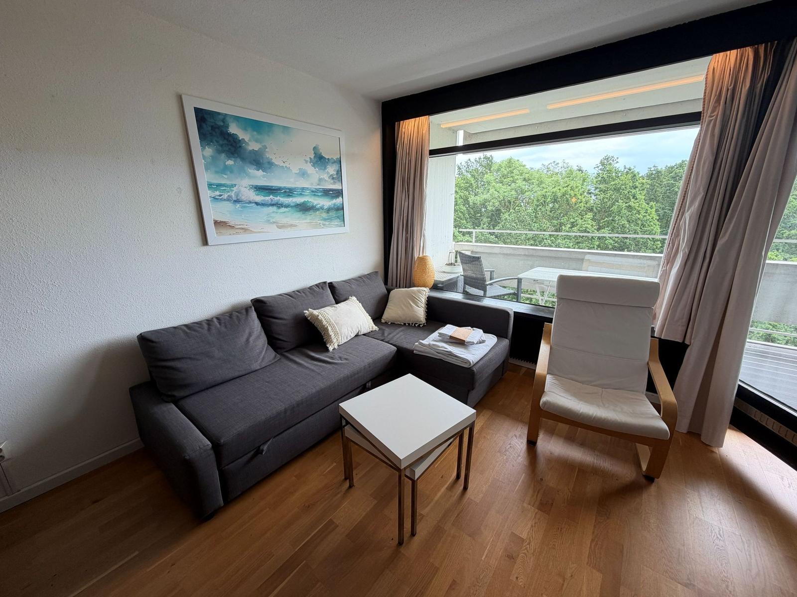 Wohnzimmer mit Sofa, Sessel und Balkon mit Blick auf Bäume.