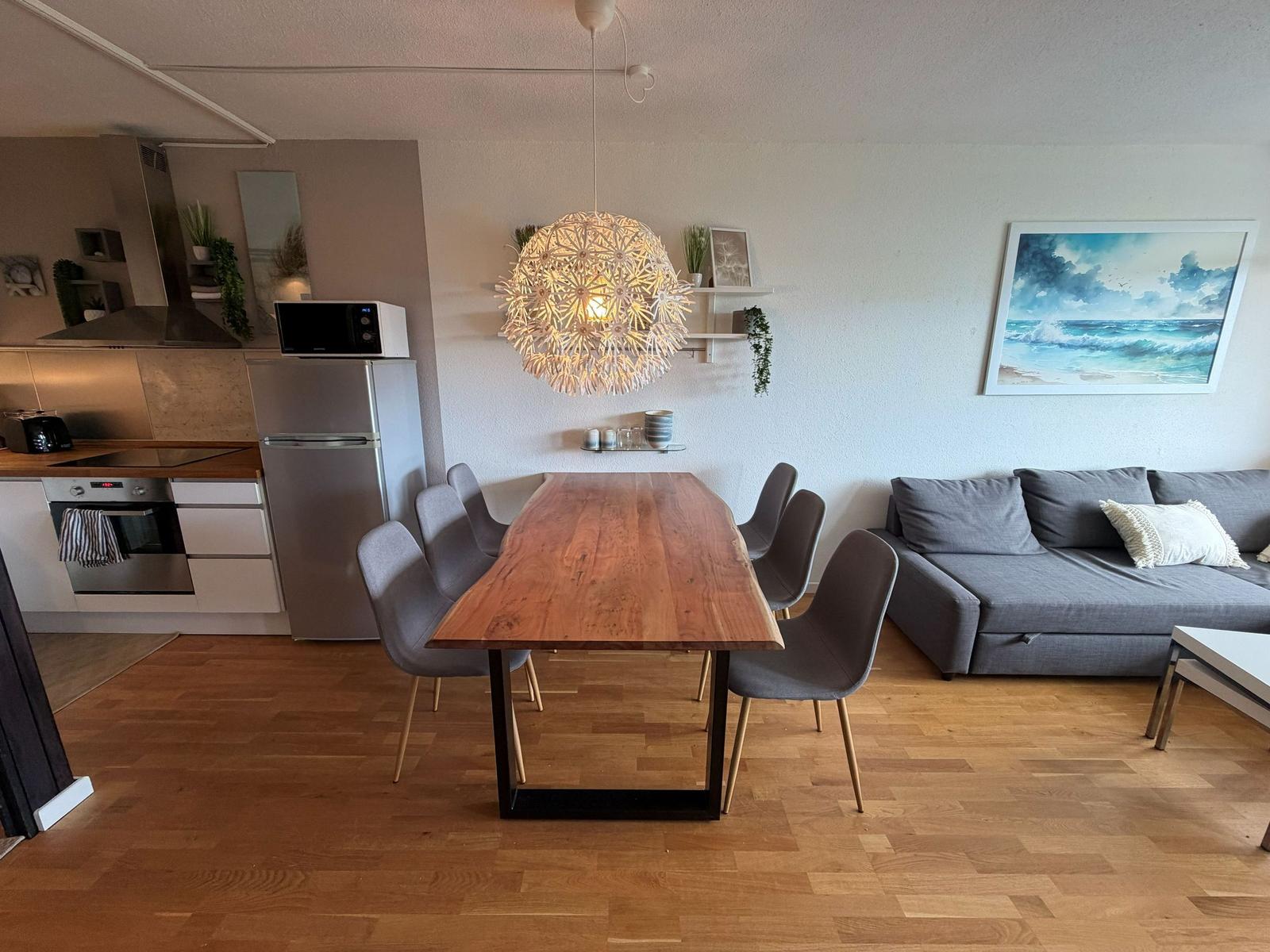 Essbereich mit Holztisch, grauen Stühlen und Sofa. Küche mit Kühlschrank und Mikrowelle.