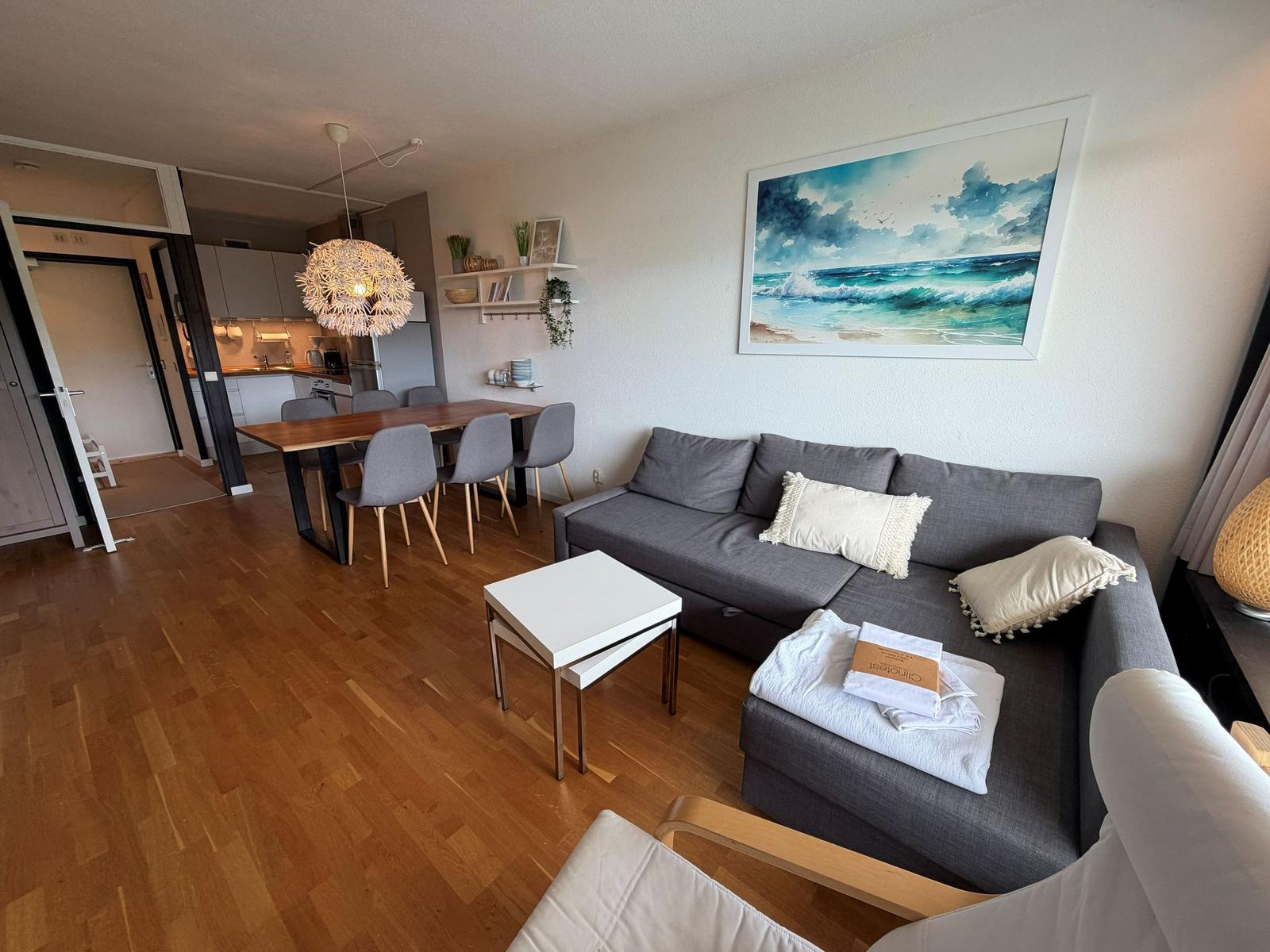 Wohnzimmer mit Sofa, Esstisch und Küchenbereich. Wandbild zeigt Meereslandschaft.