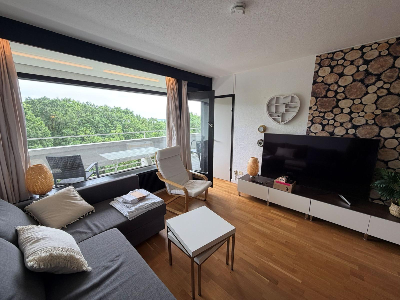 Wohnzimmer mit Sofa, Fernseher und Balkon mit Blick auf Bäume.