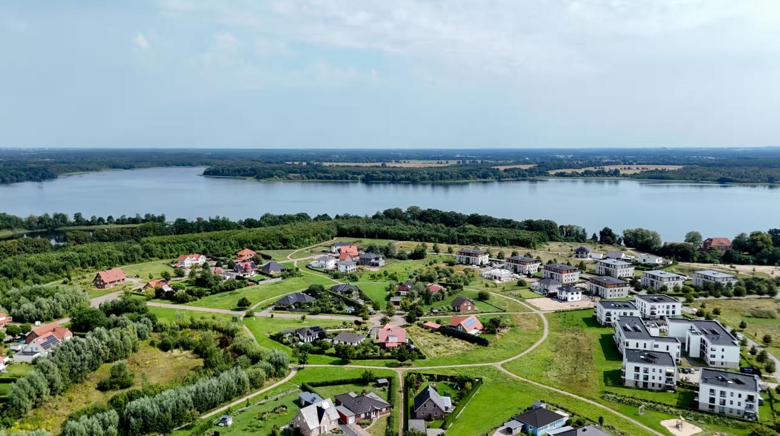 Luftbild einer Wohnsiedlung mit Grünflächen und einem großen See im Hintergrund.
