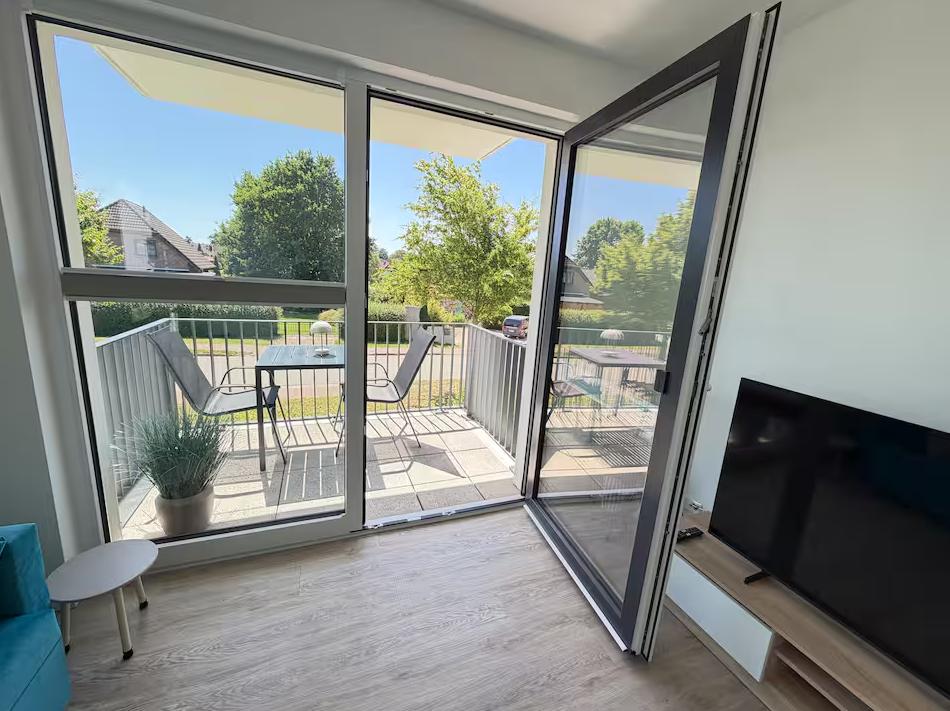 Wohnzimmer mit TV und Balkon. Balkon mit Tisch, Stühlen und Pflanze.