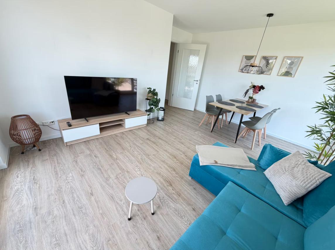 Wohn- und Essbereich mit Sofa, TV und Tisch. Holzboden und weiße Wände.