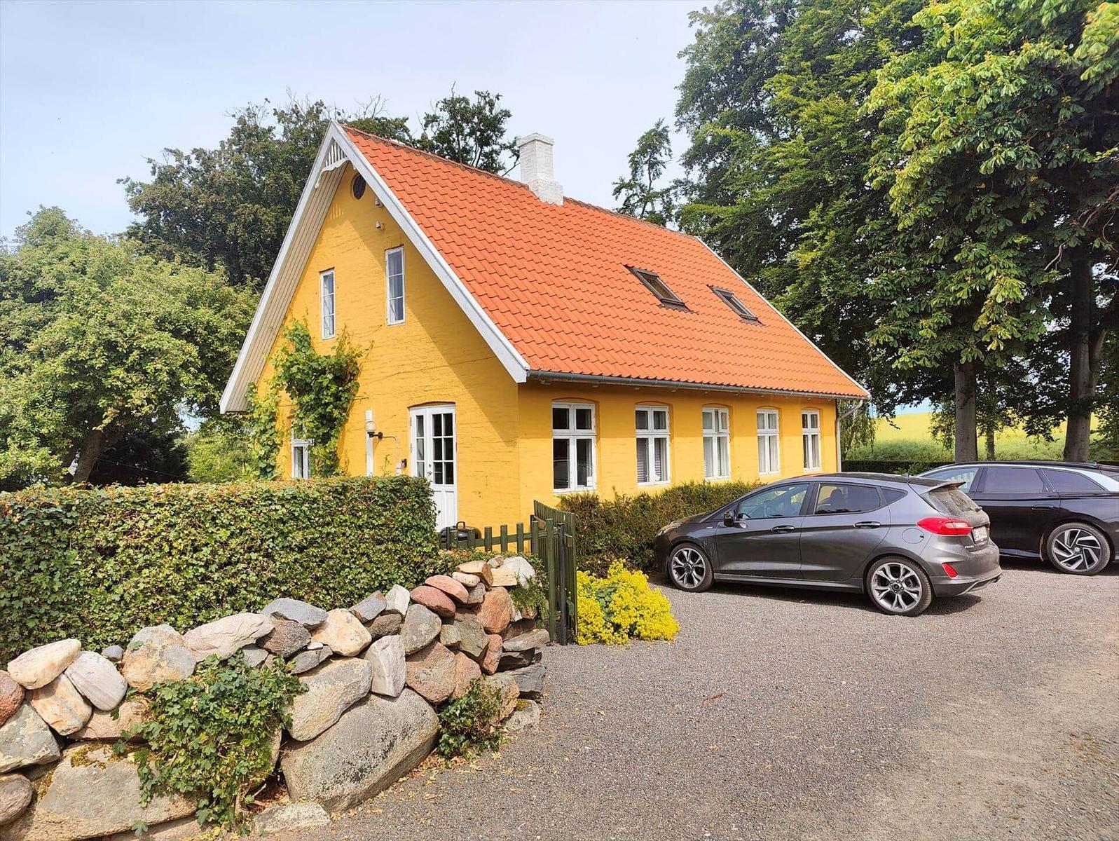 Gelbes Haus mit rotem Dach, umgeben von Bäumen und einem Steinzaun.
