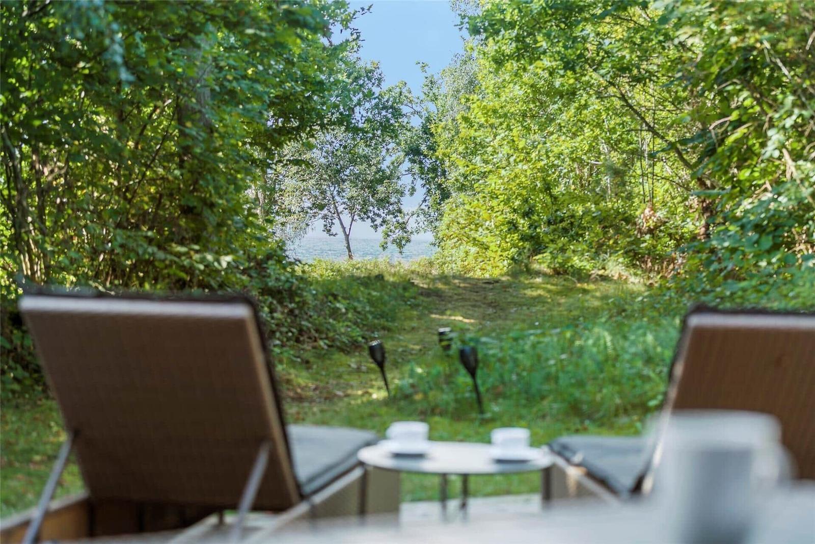 Zwei Liegestühle und Tisch auf Terrasse mit Blick durch Bäume zum See.