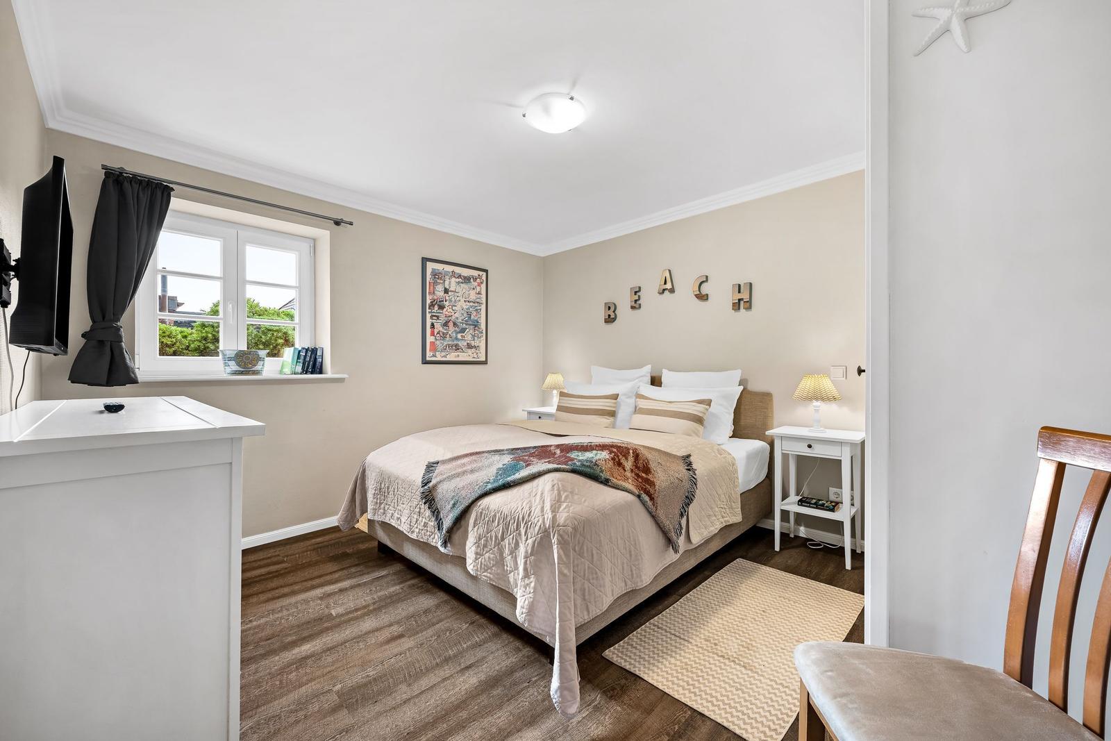 Schlafzimmer mit Bett, Nachttischen und Fenster. Wanddekoration mit BEACH.