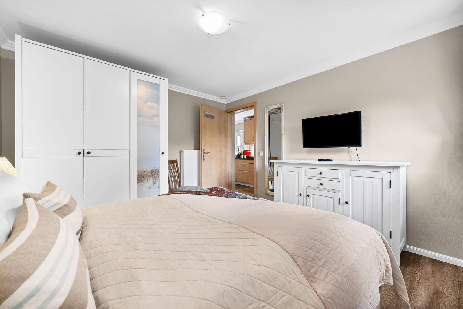Schlafzimmer mit Bett, Kleiderschrank und Fernseher auf Schrank. Tür führt in Küche.