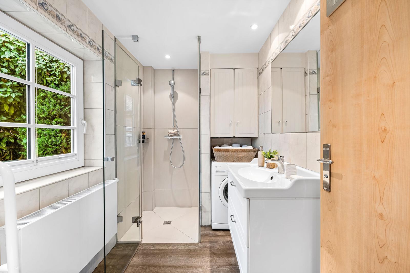 Badezimmer mit Dusche, Waschmaschine und Fenster mit Blick auf Grün.