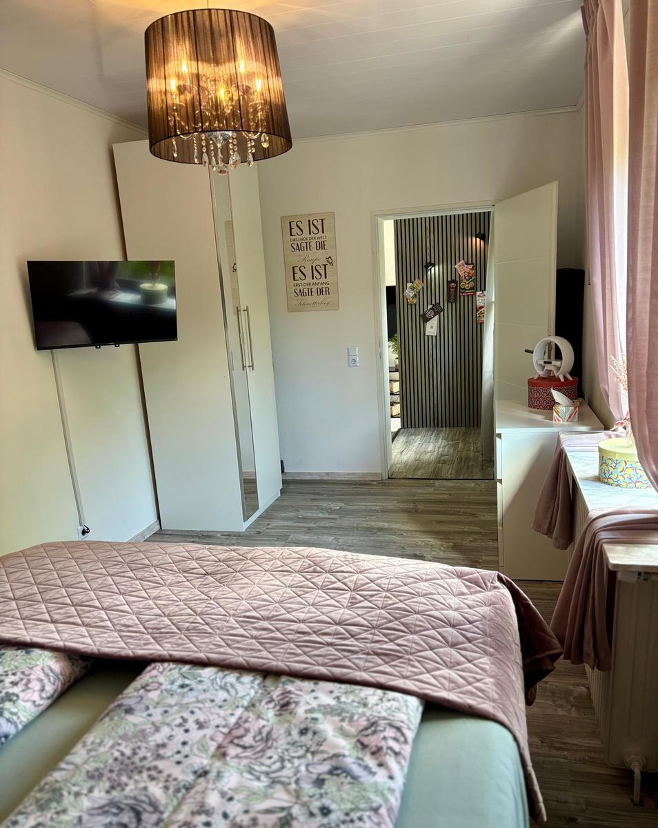 Schlafzimmer mit Bett, TV und Deckenleuchte. Tür führt in die Küche.