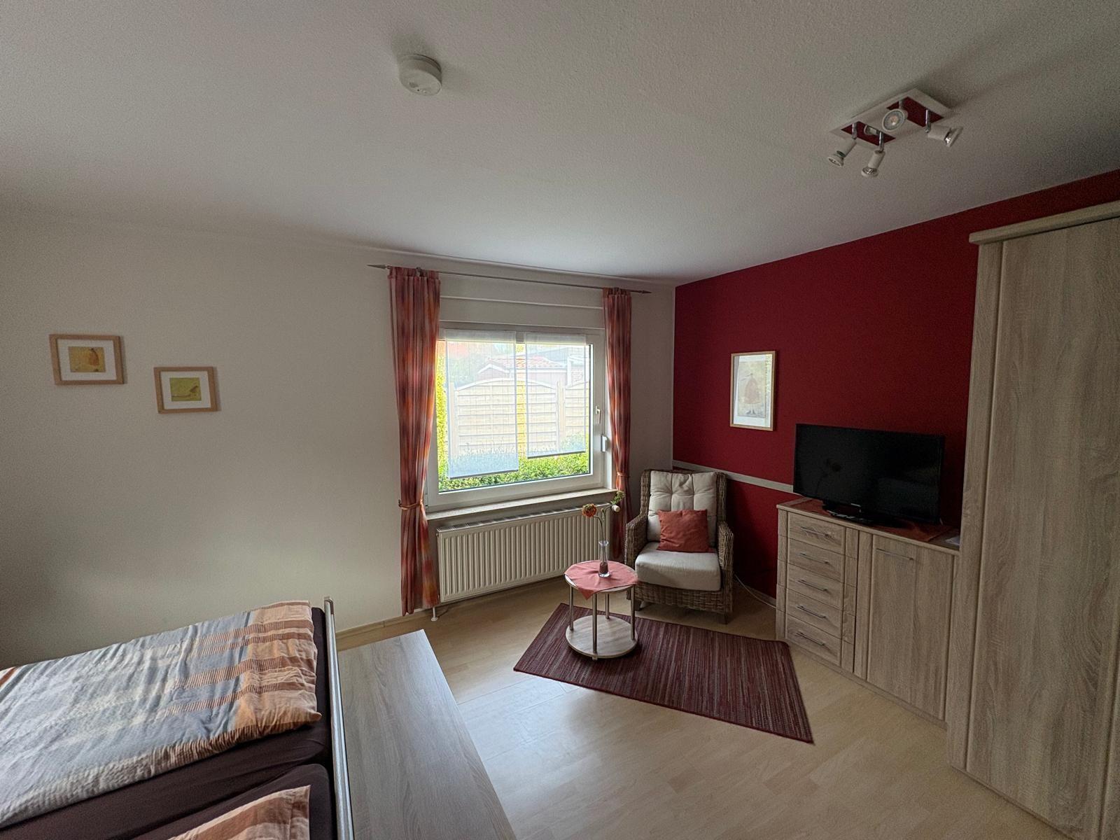 Ein Zimmer mit Bett, Sessel, Fernseher und Fenster mit Blick auf Garten.