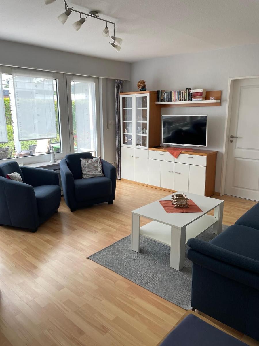Wohnzimmer mit blauen Sesseln, weißem Couchtisch und TV-Schrank. Holzboden und große Fenster.