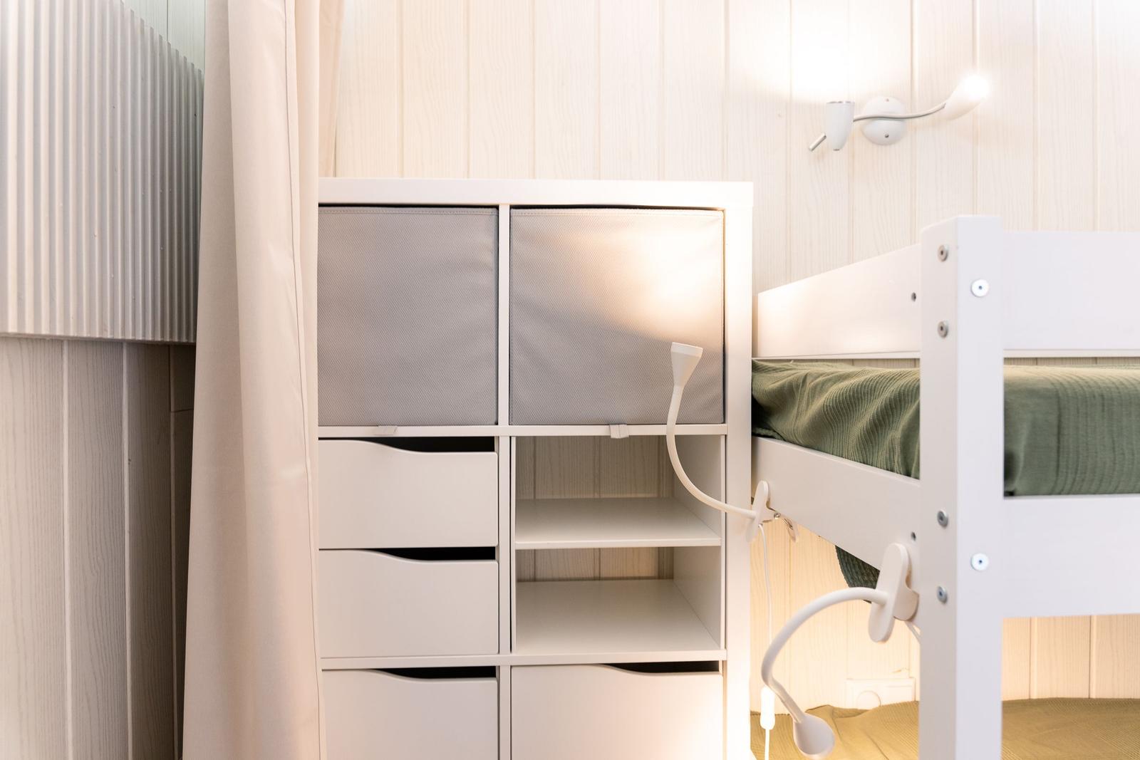 Ein weißes Bett mit grünem Bettzeug neben einem Schrank mit Fächern und einer Wandlampe.