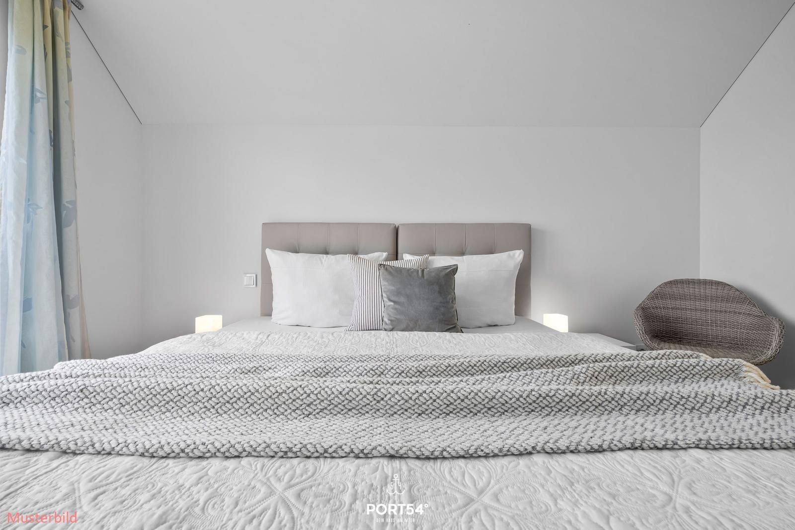 Doppelbett mit Kopfteil, Bettwäsche und Decke. Beleuchtung und Sessel sind sichtbar.