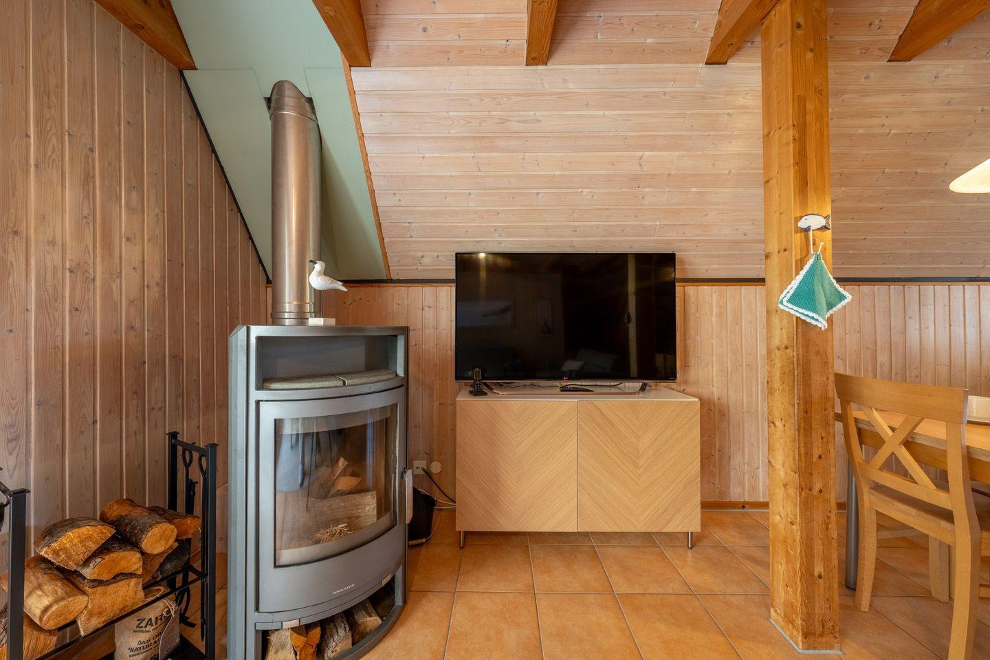 Wohnraum mit Kamin, Fernseher und Holzwand. Holzstuhl und Kaminholz sichtbar.
