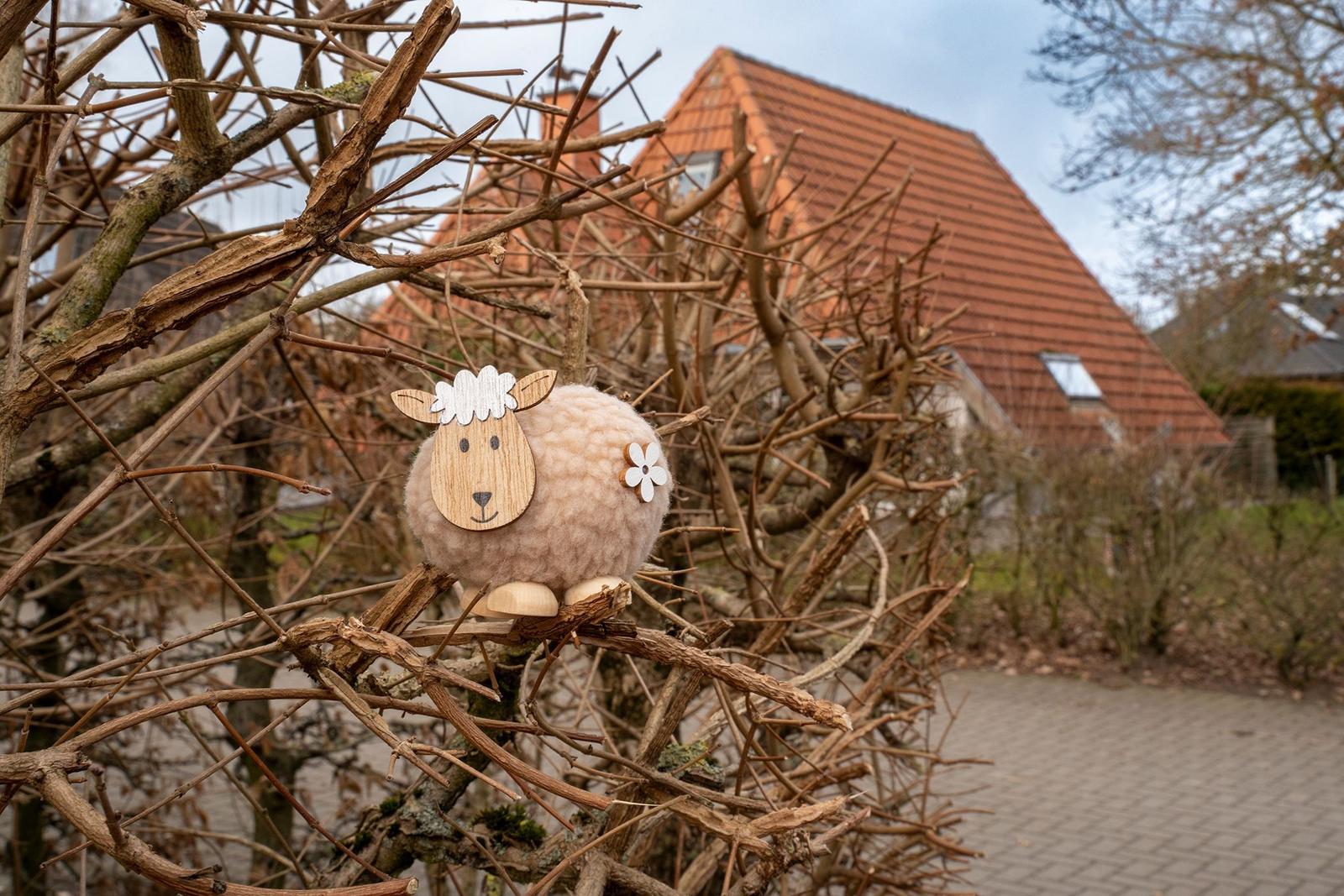 Ein hölzernes Schaf mit Wolle sitzt auf Ästen vor einem Haus mit rotem Dach.