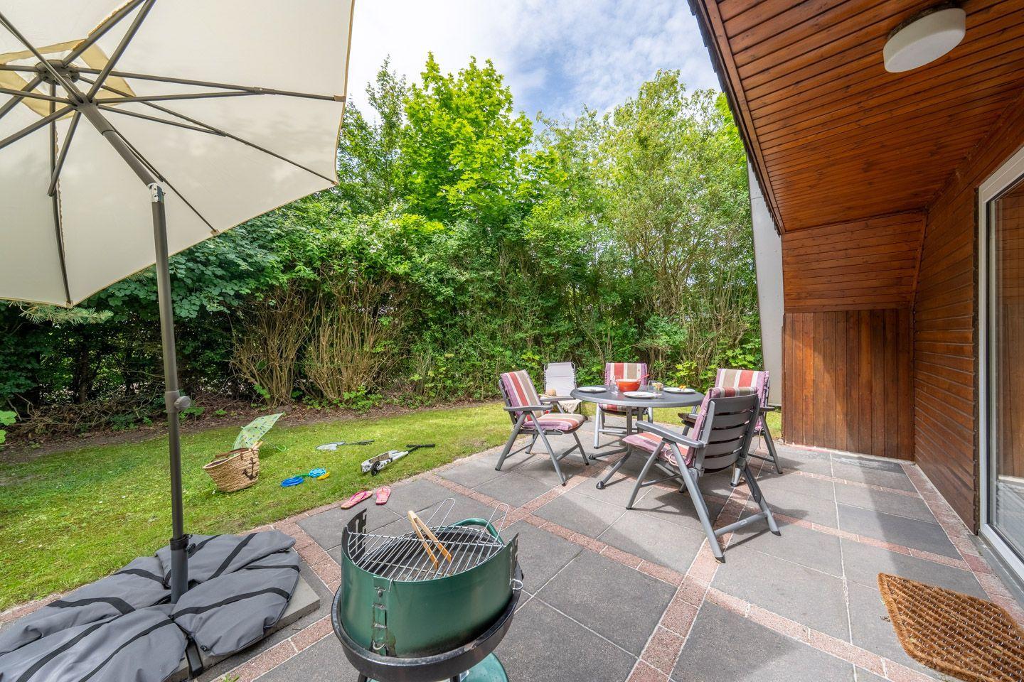 Terrasse mit Tisch, Stühlen, Grill und Sonnenschirm. Grünflächen und Bäume im Hintergrund.