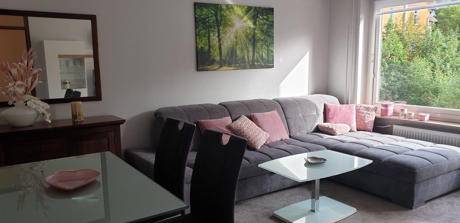 Ein Wohnzimmer mit grauem Sofa, rosa Kissen und Glas-Tisch. An der Wand hängt ein Bild.
