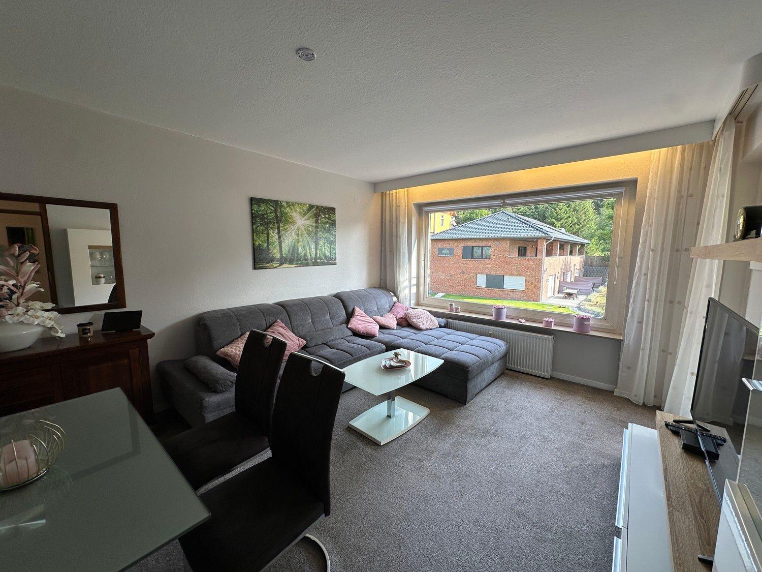 Wohnzimmer mit Sofa, Tisch und Fernseher. Großer Fensterblick auf Haus und Grünfläche.