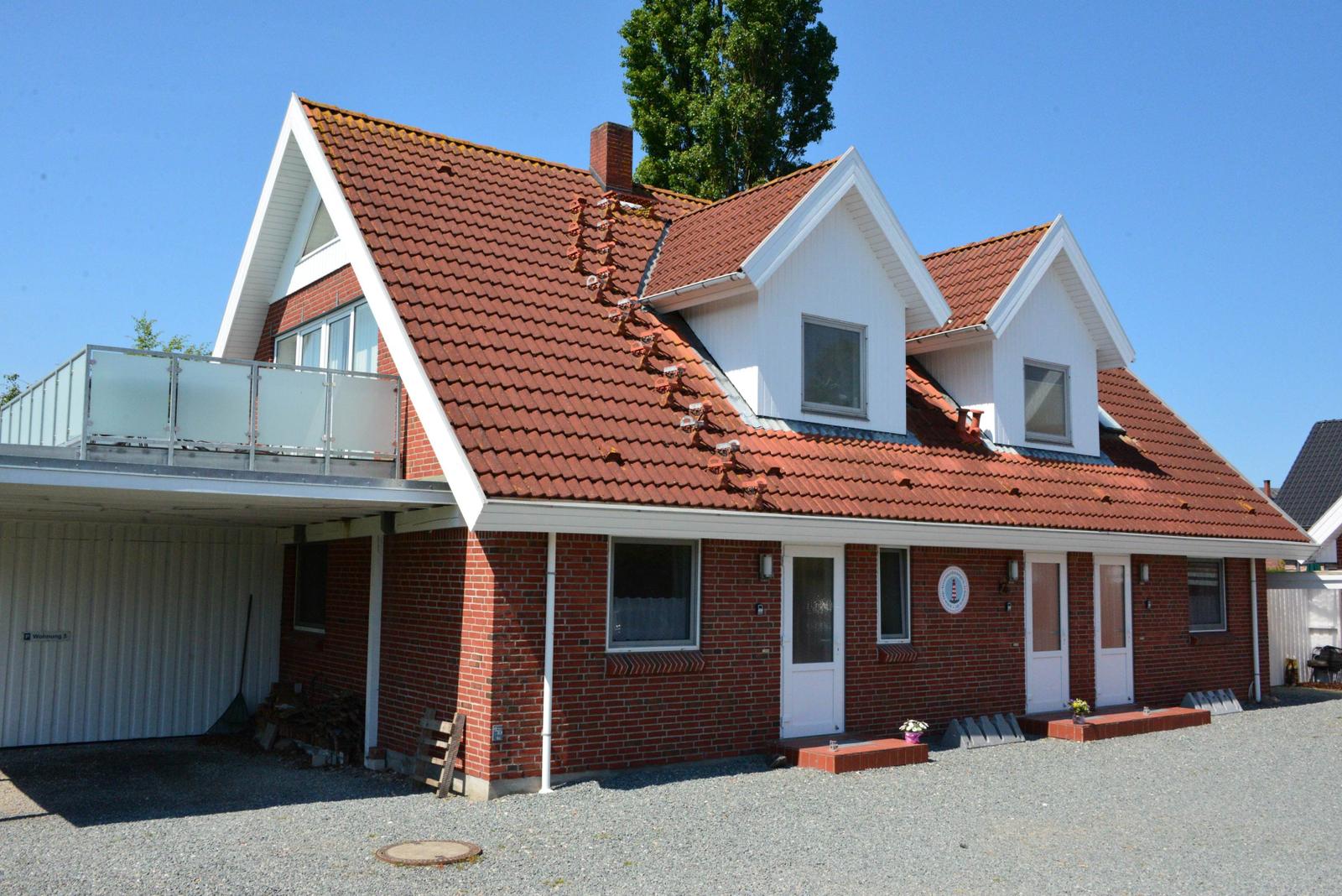 Rotes Dach, weiße Fassade, mehrere Fenster und eine Terrasse. Gravierter Boden vor dem Haus.