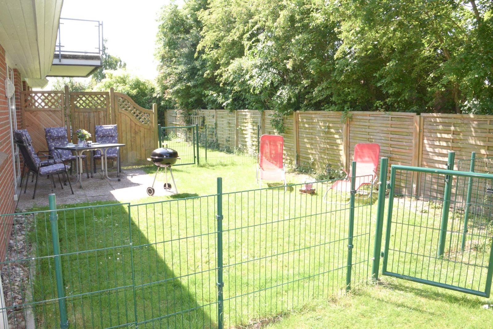 Hinterhof mit Grill, Gartenstühlen und grünem Zaun. Grasfläche und Holzgeländer im Hintergrund.