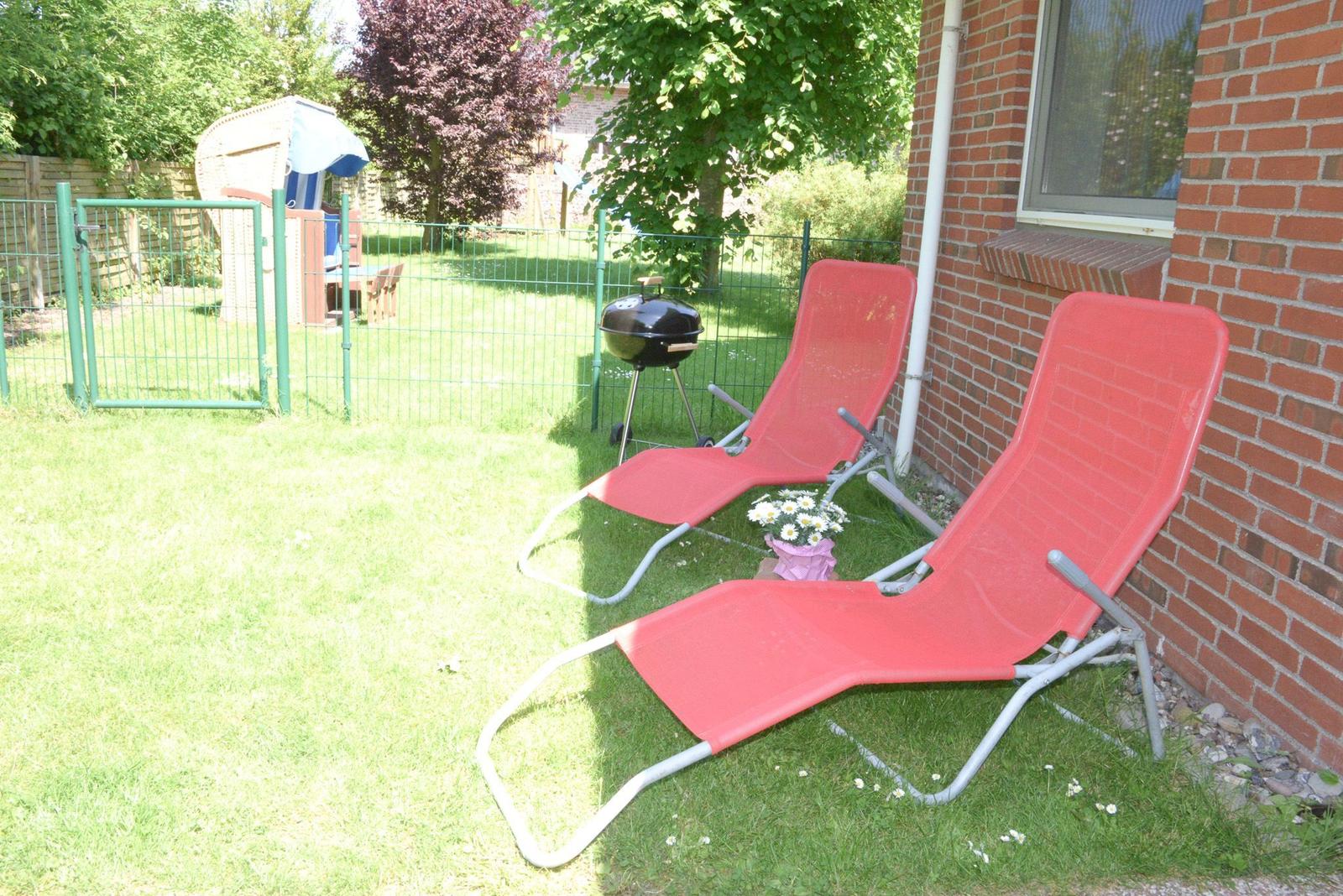 Zwei rote Liegestühle stehen im Garten neben einem Grill und einer Hauswand.