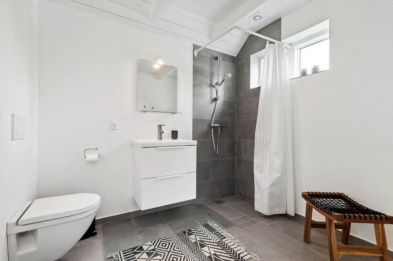 Badezimmer mit Dusche, Waschbecken und Toilette. Weiße Wände, graue Fliesen und Holzstuhl.