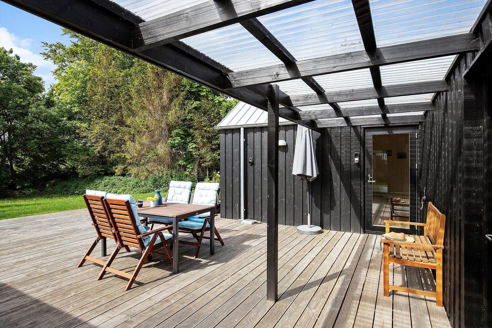 Terrasse mit Tisch, Stühlen und Sitzbank unter Dach. Hintergrund: grüner Garten und Haus.