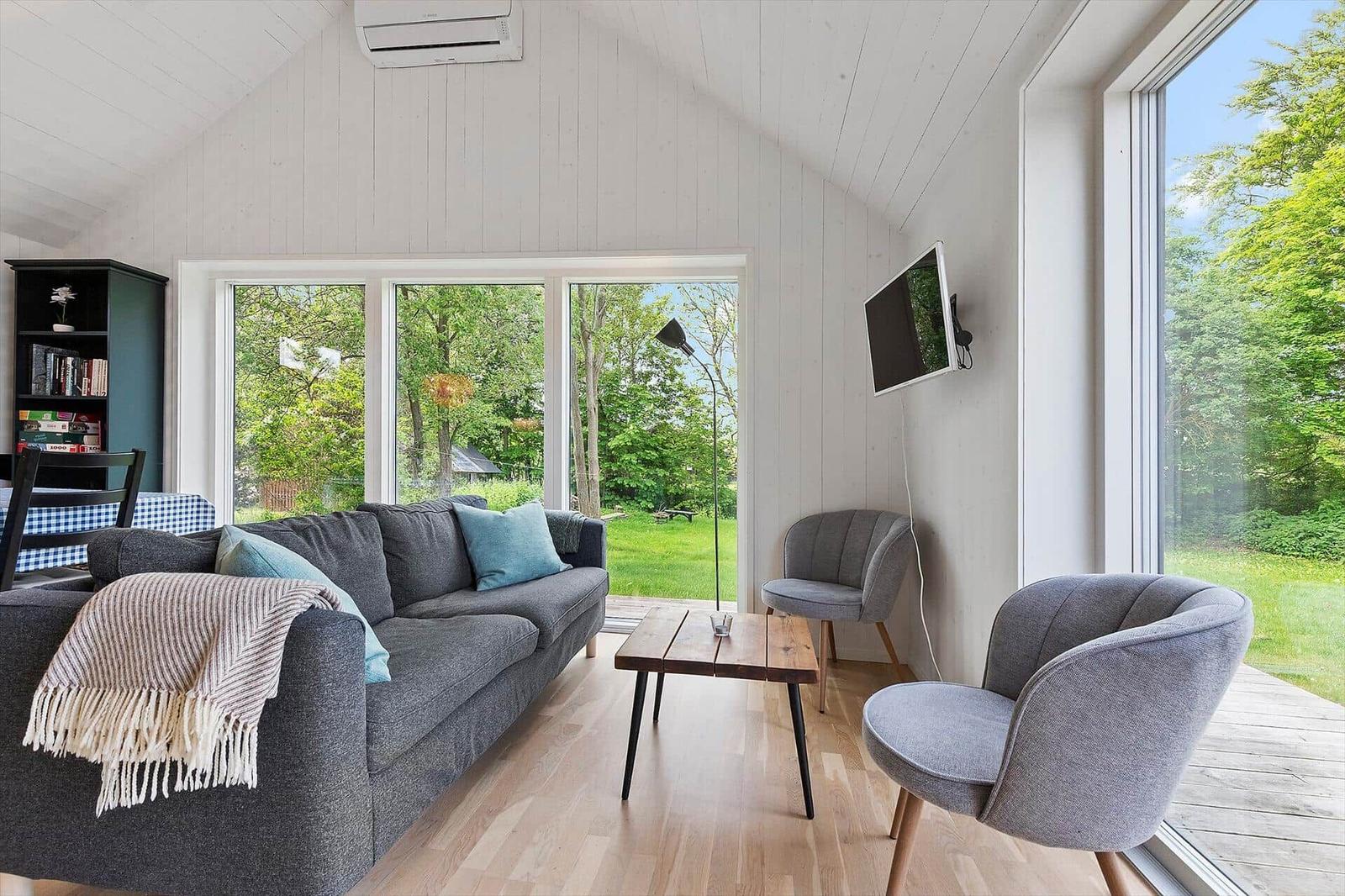 Ein Wohnzimmer mit grauem Sofa, zwei Stühlen und großen Fenstern zum Garten.