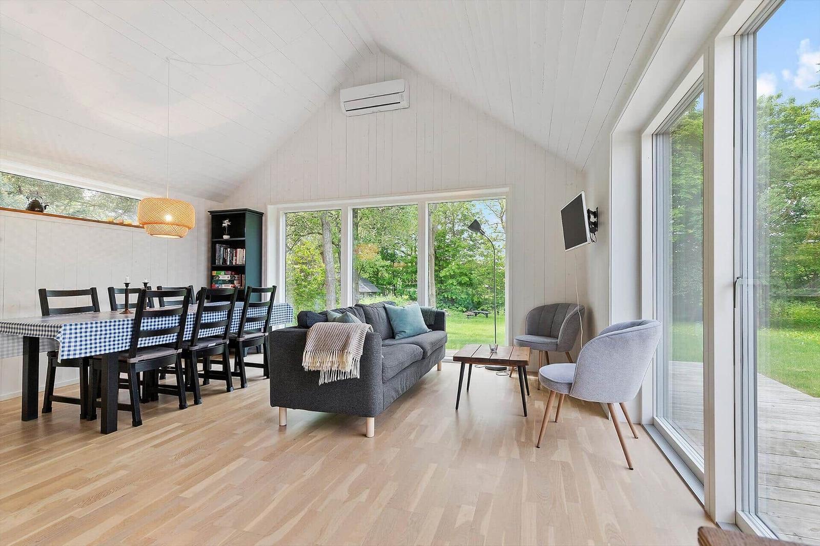 Wohn- und Essbereich mit Sofa, Tisch, Stühlen und großem Fenster. Holzboden und weiße Wände.