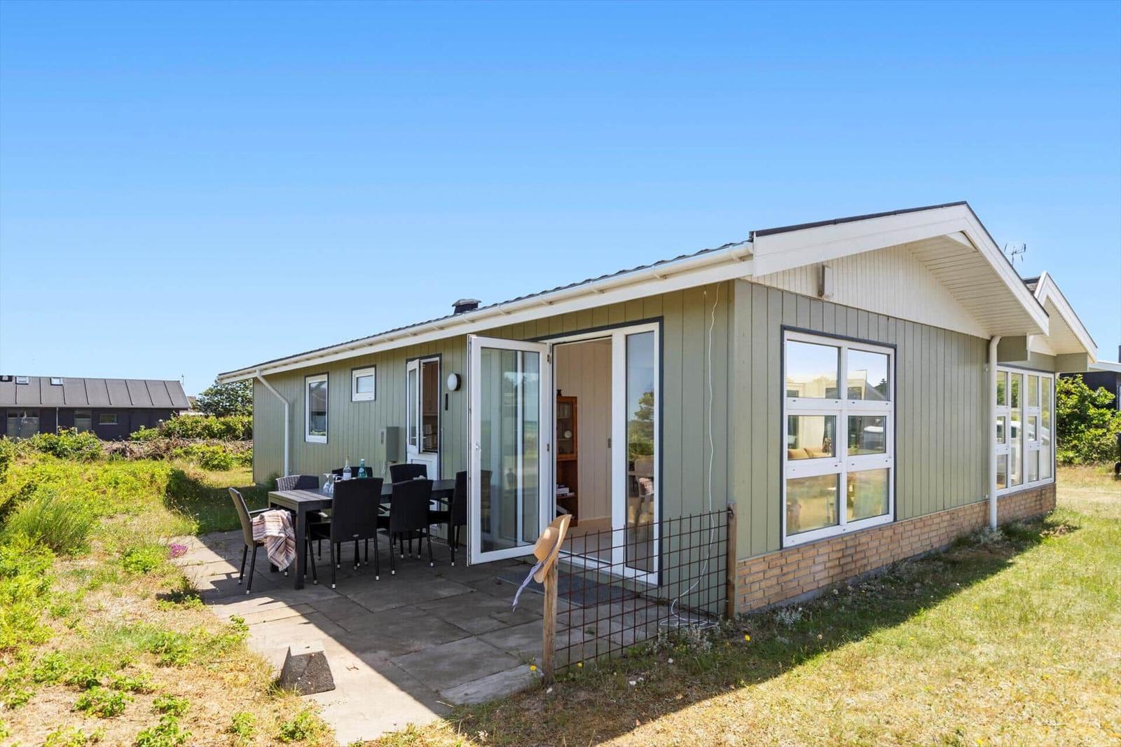 Ein Haus mit Terrasse und Gartentisch. Glasfront öffnet sich zum Innenraum.
