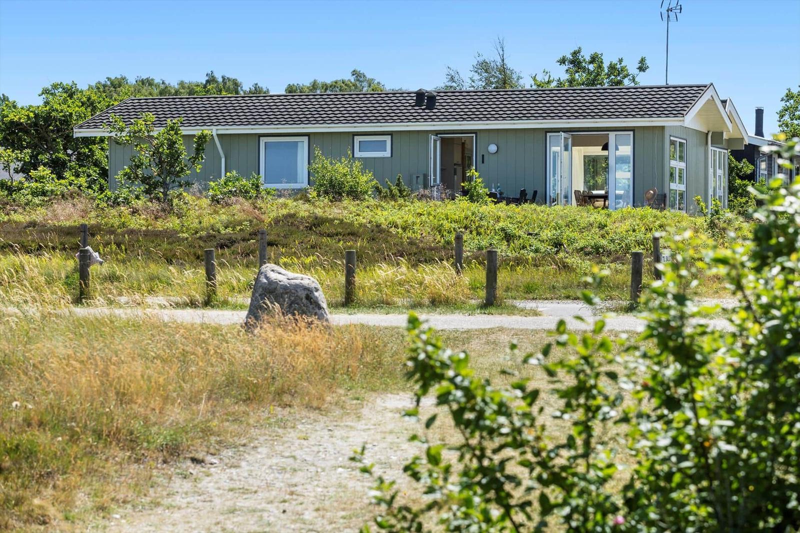 Ein graues Haus mit offenen Türen und Fenstern. Gras und Bäume umgeben es.