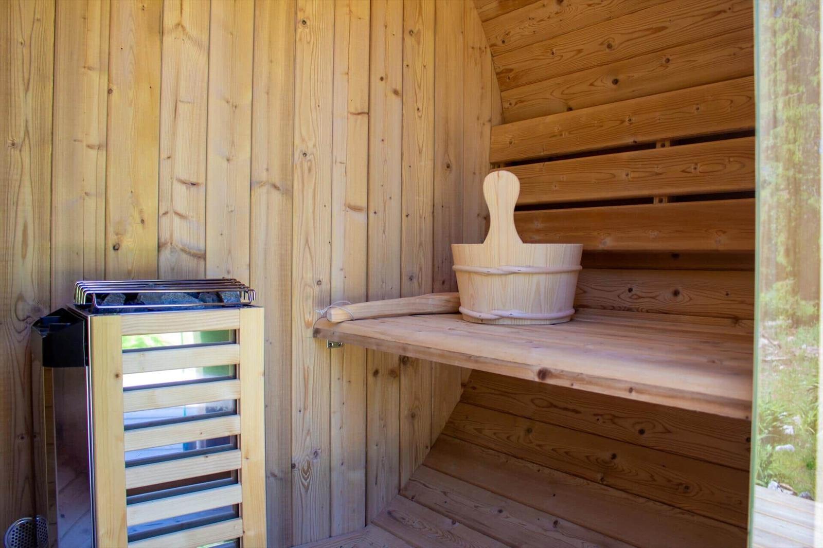 Holzsauna mit Sitzbank, Eimer und Ofen. Außenansicht durch Fenster sichtbar.