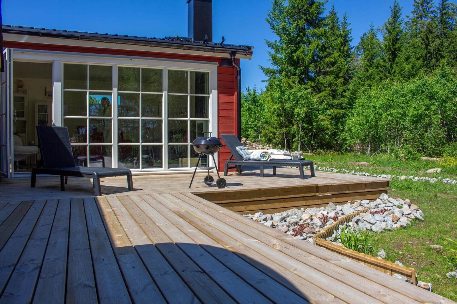 Holzterrasse mit Liegen und Grill vor einem roten Haus mit großem Fenster.