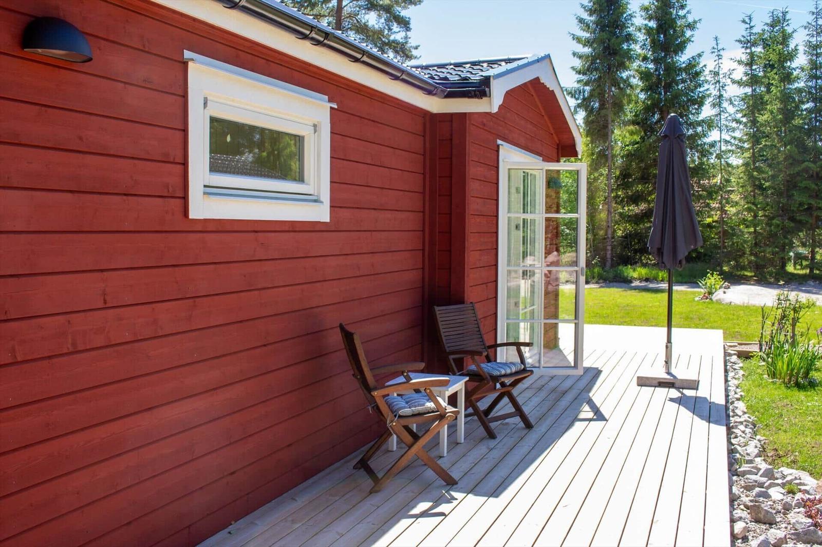 Roter Holzhaus mit Terrasse, Stühlen und Sonnenschirm. Hinterhof mit Grünfläche und Bäumen.