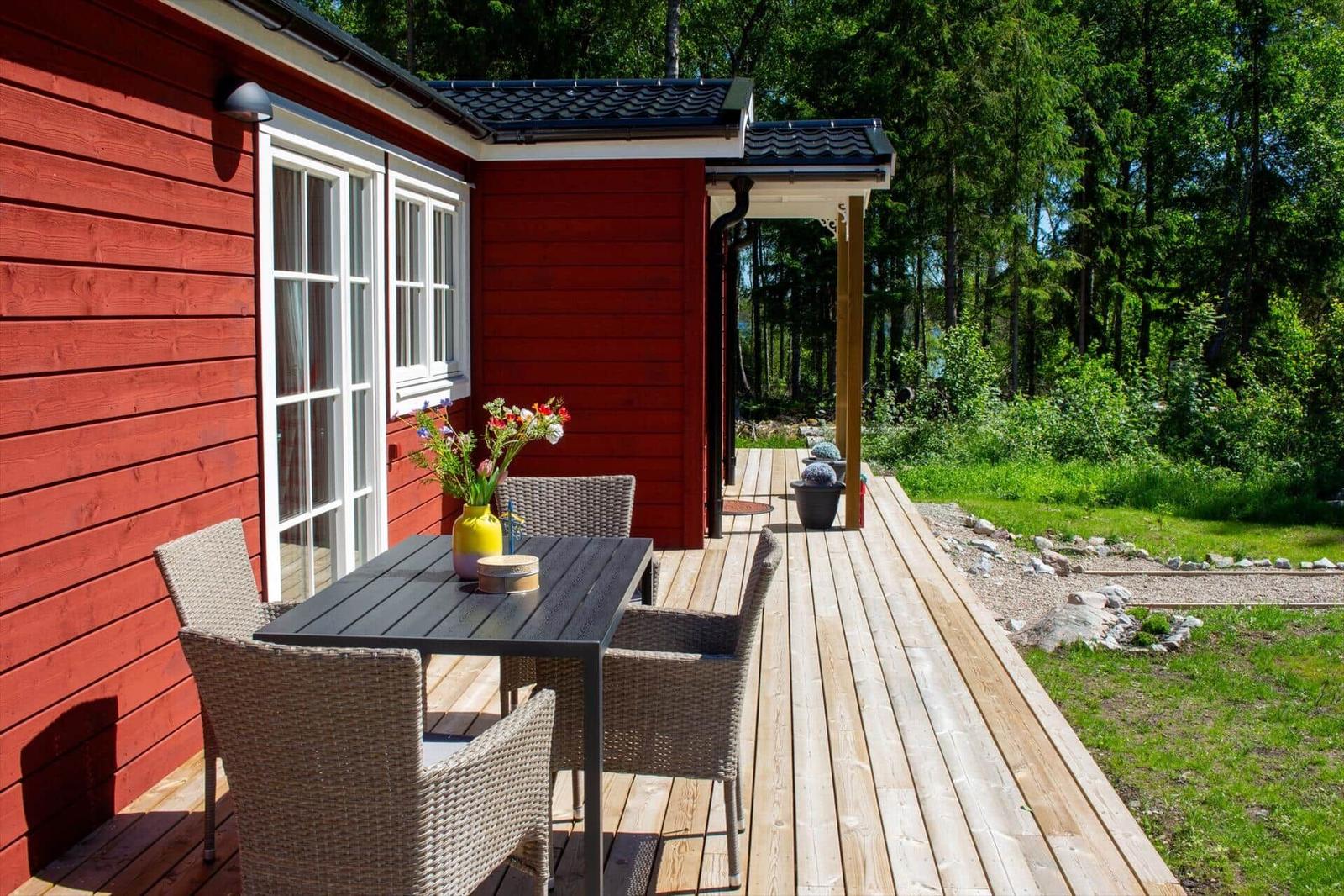 Roter Holzausbau mit Terrasse, Tisch und Stühlen. Hintergrund: Wald und Grünfläche.