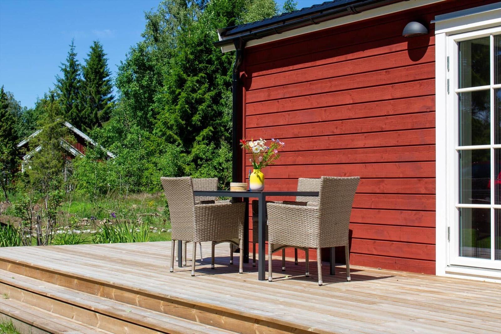 Holzterrasse mit Tisch und Stühlen vor rotem Holzhaus. Grünflächen und Bäume im Hintergrund.