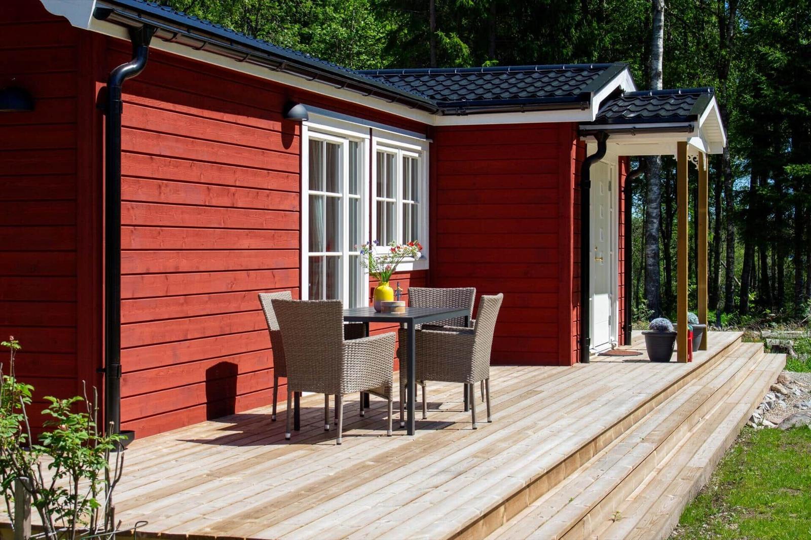 Roter Holzhaus mit Terrasse, Tisch und Stühlen, umgeben von Bäumen.